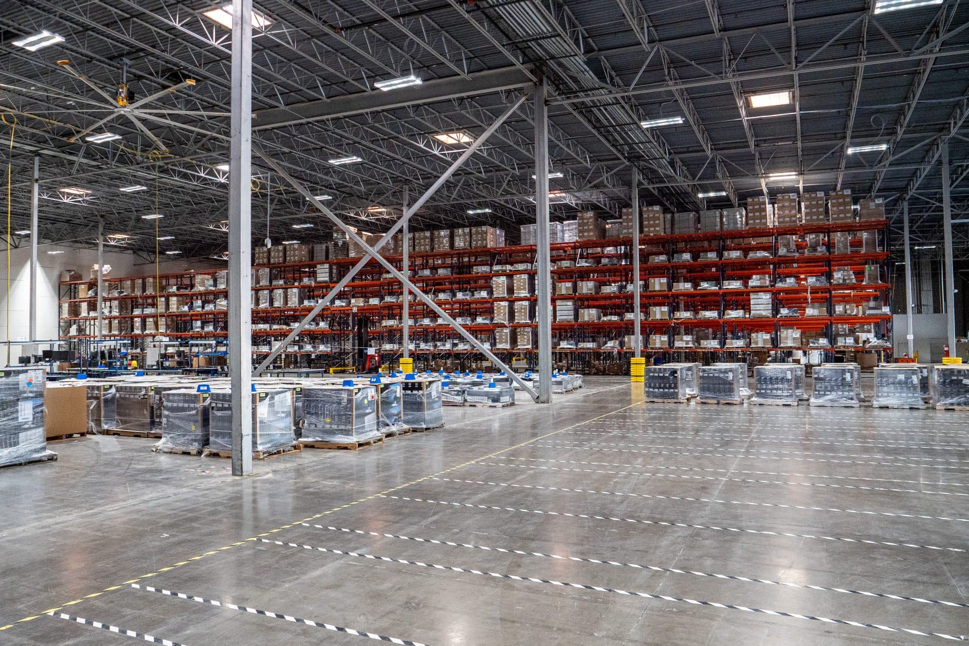 Large warehouse with high shelves stacked with boxes and palletized goods, wide aisles, and overhead lighting visible.